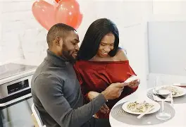 Black couple looking at a card during a romantic dinner with red heart balloons.