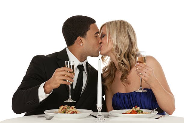 Couple in formal wear kissing over a candlelit dinner with champagne flutes and pasta.