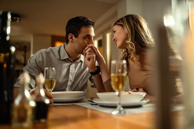 Man affectionately kisses a woman's hand during a romantic dinner with wine and food.