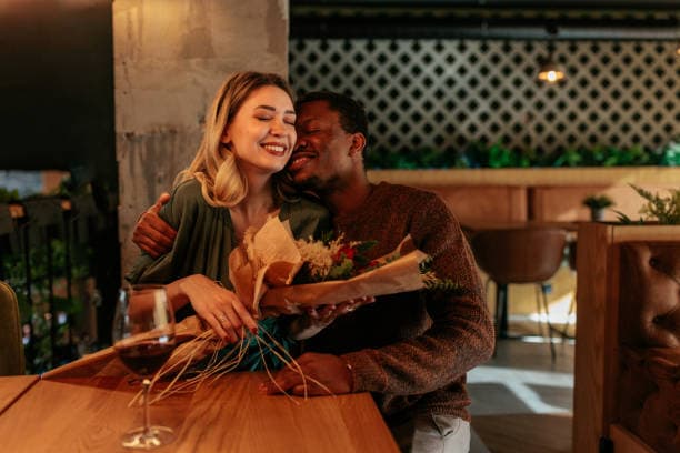 Smiling interracial couple embracing at a restaurant table while holding a bouquet of flowers.