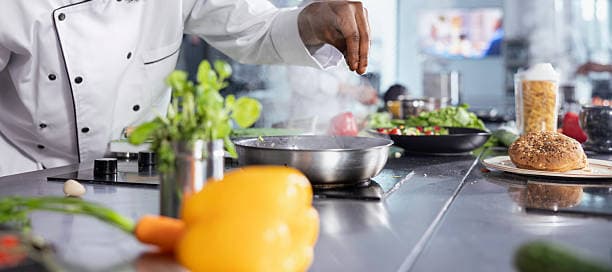 Professional chef seasoning food in a pan on a stove surrounded by fresh ingredients.