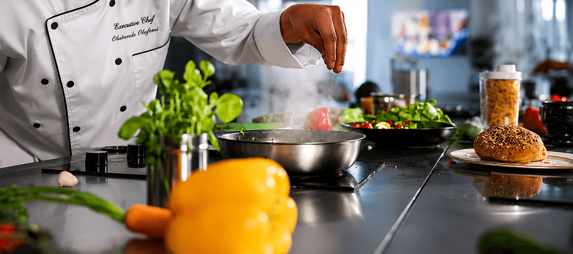 Executive chef seasoning food in a pan with fresh vegetables and ingredients on a counter.
