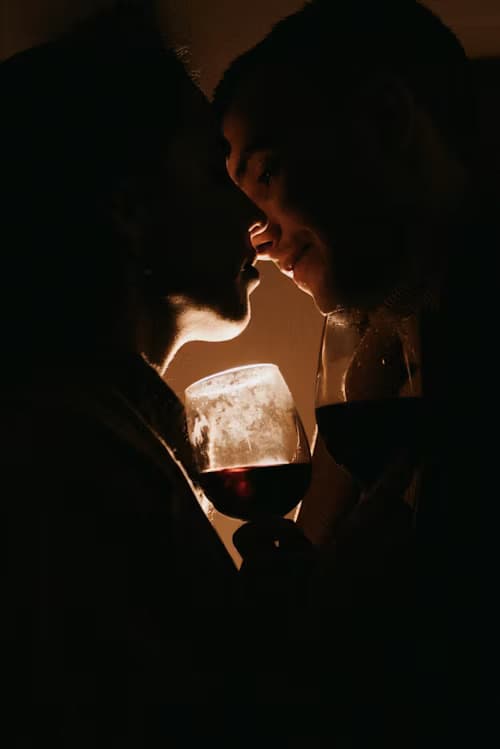 Silhouette of a couple leaning in for a kiss, holding wine glasses in warm light.